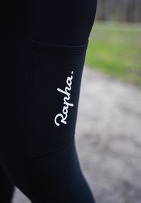 Leggings neri realizzati in materiale elastico con una tasca laterale e il logo bianco "Rapha." sulla coscia. Texture liscia, design aderente.
