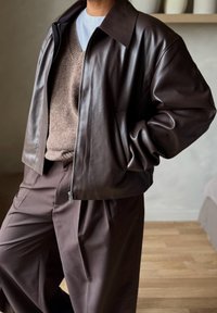 Άτομο που φοράει καφέ δερμάτινο μπουφάν πάνω από μπεζ πουλόβερ με V λαιμόκοψη και καφέ πλισέ παντελόνια, όρθιο σε εσωτερικό χώρο με το ένα χέρι στην τσέπη.