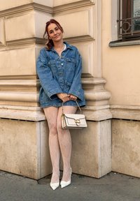 Džínová bunda cez biely top; vzorované sieťované pančuchy; biele špicaté podpätky; krémová kabelka so zlatými doplnkami; stojí pri kamennej stene.