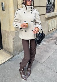 Jasnoszary płaszcz dwurzędowy z wysokim kołnierzem, czarne guziki. Brązowe dopasowane spodnie i brązowe spiczaste buty. W ręku czarna torebka.