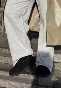 Personne portant un pantalon de survêtement gris, des chaussures noires sans lacets et un manteau beige brillant, debout sur des marches en pierre sous le soleil.