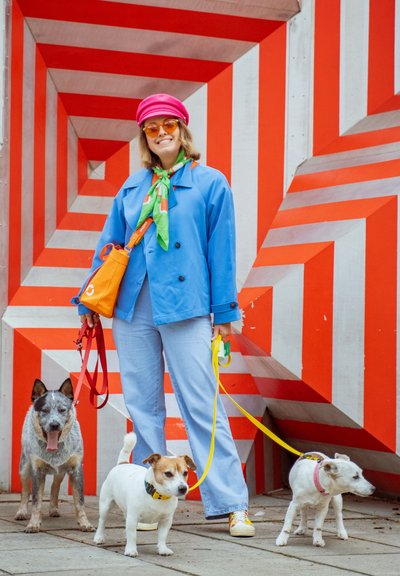 Mujer con ropa llamativa sosteniendo las correas de tres perros, de pie frente a una pared con un patrón geométrico rojo y blanco.