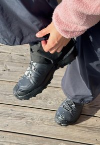 Personne portant des bottes de neige noires et un pantalon foncé, ajustant l'ourlet du pantalon par-dessus la botte sur une terrasse en bois avec une fine couche de neige.