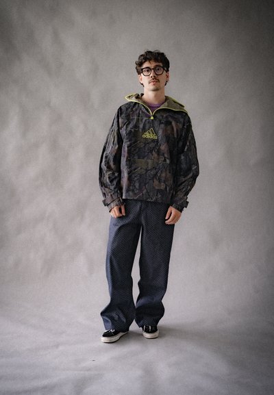 Rompevientos de patrón camouflage con una cremallera y logotipo amarillos, combinado con pantalones oscuros de patrón y zapatillas negras. Ajuste texturizado y casual.