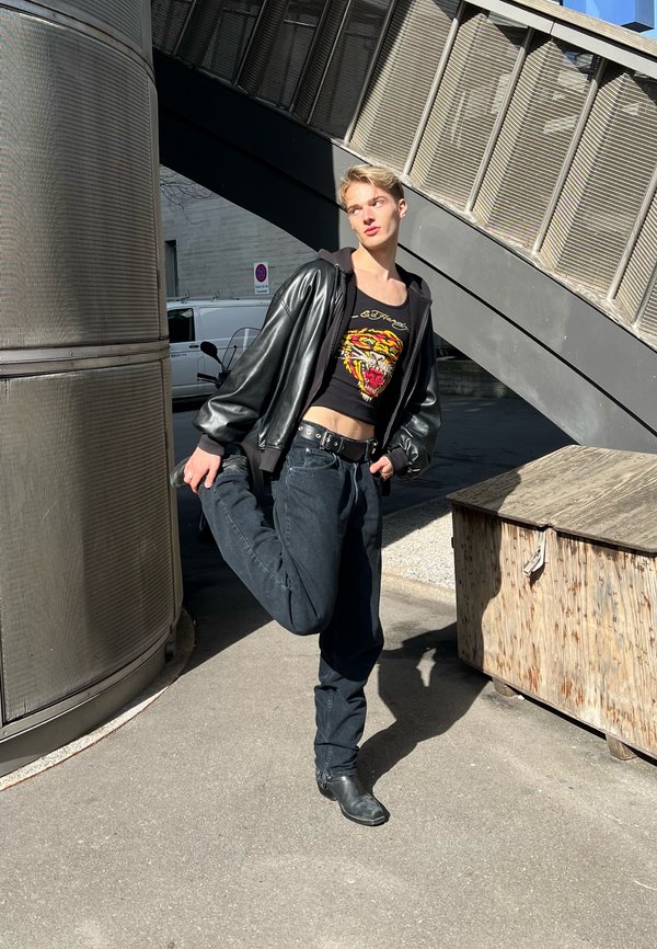 Junge Person in schwarzer Lederjacke und Tiger-Print-Top streckt ein Bein nach hinten und steht auf dem städtischen Gehweg unter einer Metalltreppe.