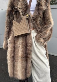 Personne portant un long manteau en fourrure marron sur un pantalon blanc, tenant un sac à main marron à motifs avec deux poignées et une bandoulière.