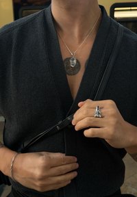 Black knitted top with a deep V-neck, layered silver chain necklace featuring a clear stone pendant, silver ring with a geometric design, and black bag strap.