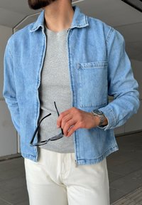 Chaqueta de mezclilla de color azul claro con un bolsillo en el pecho, camiseta gris de canalé debajo y pantalones blancos. Mano sosteniendo gafas negras, llevando un reloj de plata.