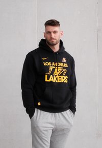 Zwarte hoodie met het "Los Angeles Lakers" logo in geel, voorzien van een kangoeroezak en een capuchon met koord, gecombineerd met grijze joggingbroeken.
