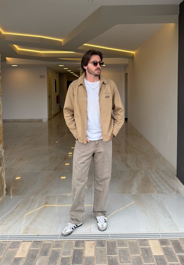 Tan jacket over a white shirt, grey trousers, and grey trainers. Standing on a polished floor beneath linear ceiling lights in a bright corridor.