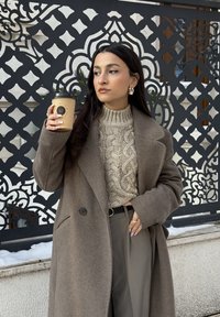 Γυναίκα με μπεζ πουλόβερ πλεξούδα και μακρύ καφέ παλτό κρατάει κούπα καφέ, στέκεται μπροστά σε διακοσμητική μαύρη και άσπρη μεταλλική οθόνη με σχέδια.