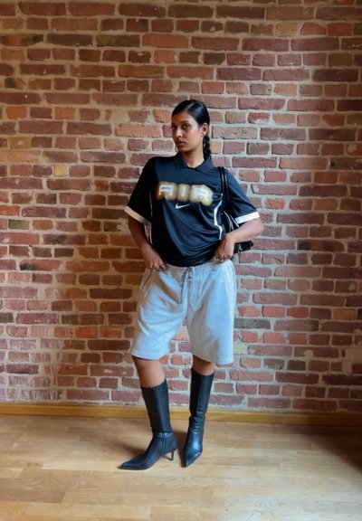 Camisa negra de manga corta con el texto "AIR" en dorado y acentos en blanco, combinada con pantalones cortos grises y botas negras hasta la rodilla sobre un suelo de madera.