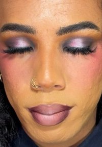Primo piano di un volto con fondotinta sfumato, labbra scure e ombretto viola con scintillio. Le ciglia sono lunghe e c'è un anello al naso.