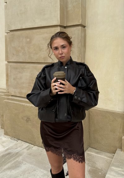 Chaqueta de cuero texturizado negra con un alto cuello, superpuesta a una falda marrón con ribete de encaje. Sosteniendo una taza de café, de pie frente a un fondo de piedra.