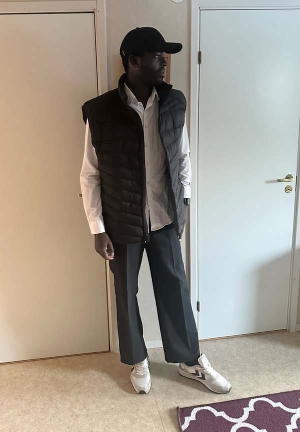 Άνδρας με μαύρο καπέλο, μαύρο αμάνικο μπουφάν, λευκό πουκάμισο, σκούρο παντελόνι και λευκά αθλητικά παπούτσια στέκεται μέσα σε εσωτερικό χώρο μεταξύ δύο κλειστών πορτών.