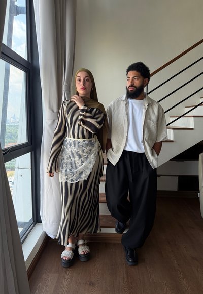 Mujer con vestido estampado y hiyab está junto a la ventana; hombre con barba, camisa clara y pantalones negros está en las escaleras dentro de la casa.
