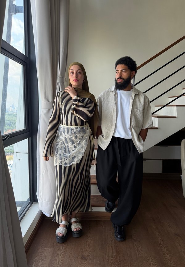 Moteris dėvinti raštuotą suknelę ir hidžabą stovi prie lango; vyras su barzda ir šviesia marškinėliais bei juodos spalvos kelnėmis stovi laiptinėse viduje.