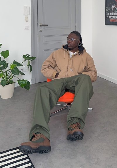 Hombre con gafas y cabello trenzado, vestido con chaqueta marrón y pantalones verdes, sentado en una silla naranja en una habitación minimalista con planta y puerta gris.