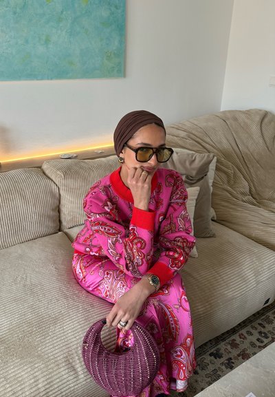 Mujer con pañuelo marrón en la cabeza, gafas de sol grandes y un conjunto floral rosa, sentada en un sofá beige sosteniendo un bolso trenzado morado.