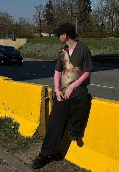 Persona sentada en una barrera amarilla, vistiendo una camisa de botones de manga corta con un estampado fotográfico y pantalones sueltos negros. Zapatillas negras.