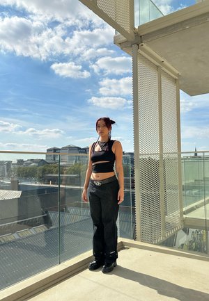 Top sin mangas negro de corte corto, jeans oscuros de cintura alta y zapatos de plataforma negros gruesos. De pie en un balcón con barandilla de cristal y cielo nublado.