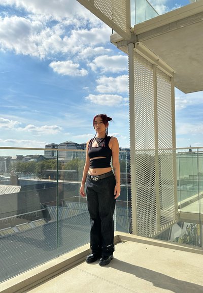 Top sin mangas negro de corte corto, jeans oscuros de cintura alta y zapatos de plataforma negros gruesos. De pie en un balcón con barandilla de cristal y cielo nublado.