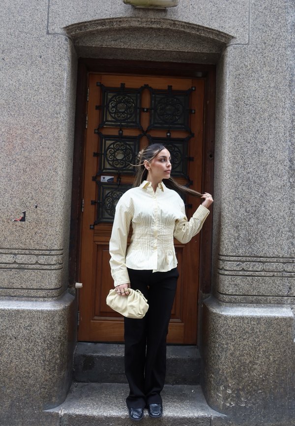 Femeie cu părul tras înapoi poartă o bluză bej în dungi și pantaloni negri, ținând o poșetă bej, stând în fața unei uși de lemn.