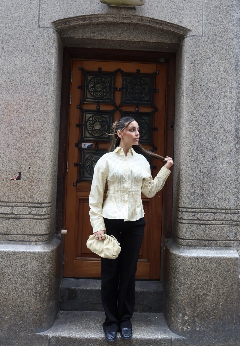 Frau mit zurückgebundenem Haar trägt eine gestreifte beige Bluse und schwarze Hose, hält eine beige Handtasche und steht vor einer Holztür.