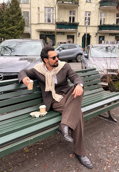 Hombre con traje marrón y bufanda crema sentado en un banco verde sosteniendo una taza de café, con coches estacionados y edificios al fondo.