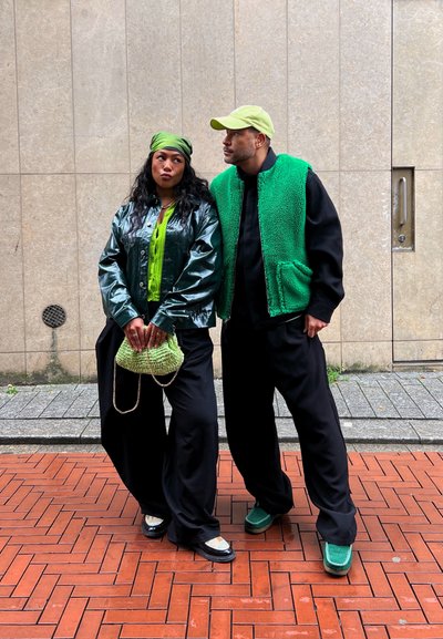 Una chaqueta de vinilo verde brillante sobre una blusa verde brillante, combinada con pantalones baggy negros. Los accesorios incluyen un bolso texturizado verde y unos zapatos a juego.