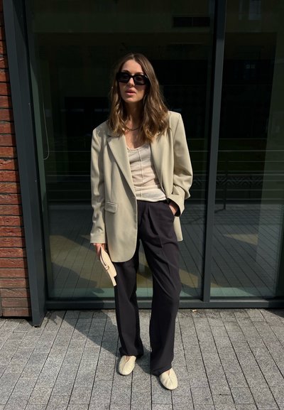 Mujer vestida con blazer beige, top blanco, pantalones negros y zapatos beige, está de pie al aire libre sosteniendo un pequeño bolso beige, lleva gafas de sol.