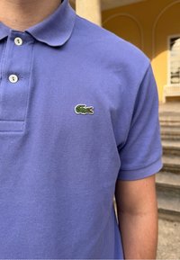 Vijolična polo majica iz teksturirane tkanine s ovratnikom in tremi gumbi, ki na levem prsnem delu prikazuje majhen zeleni krokodil.