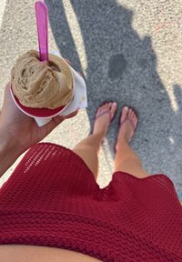 Coppa di gelato con una pallina di colore marrone chiaro e un cucchiaino rosa, tenuta in mano. Indumenti rossi lavorati a maglia e piedi in ciabatte viola sottostanti.