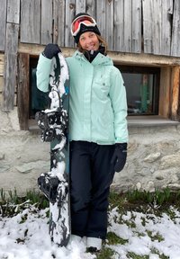 Ένας σνόουμπορντερ με ένα ανοιχτό μέντα μπουφάν και μαύρα παντελόνια κρατάει ένα σνόουμπορντ με δέστρες, περιτριγυρισμένος από χιόνι και πράσινα φυτά.