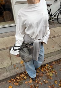 Pelēka ādas soma ar rāvējslēdzēju un rokturi, nēsāta pār gaiši pelēku džemperi, kas sasiets virs baltas garroku krekla un brīvām zilām džinsu biksēm.