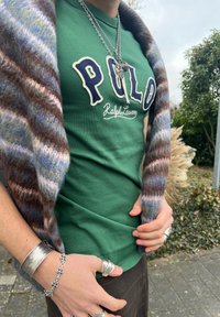 Personne portant un polo vert Ralph Lauren, des colliers superposés, des bagues et des bracelets en argent, tenant une écharpe en laine rayée en plein air.