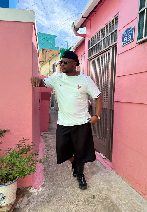 Homme portant des lunettes de soleil, un béret noir, un t-shirt de sport blanc et un short noir s’appuie contre un mur rose vif dans une ruelle étroite extérieure avec des plantes.