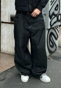 Personne portant un jean noir surdimensionné, des baskets blanches et une veste noire, debout sur un trottoir de la ville près d'un mur couvert de graffiti.