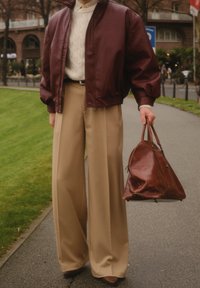 Veste bomber en cuir bordeaux portée sur un pull en maille torsadée crème, associée à un pantalon large beige et un sac en cuir marron.
