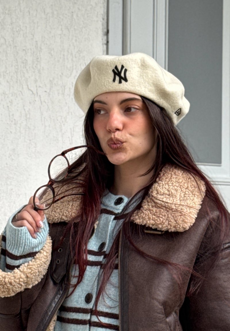 Młoda kobieta w beżowym berecie z napisem NY i brązowej kurtce z owczej skóry trzyma okulary przy ustach, patrząc na bok w pomieszczeniu.