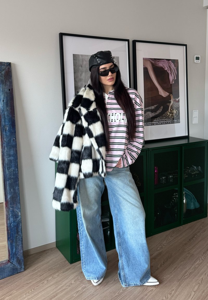 Femme portant un manteau en fourrure à carreaux noir et blanc sur l'épaule, une chemise rose rayée, un jean large bleu clair, une casquette en cuir noire et des lunettes de soleil à l'intérieur.
