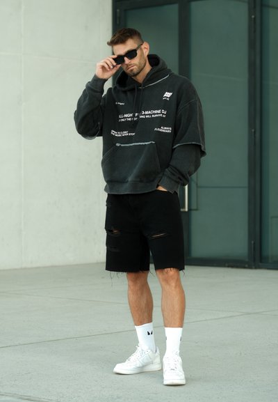 Sudadera negra de gran tamaño con gráficos de texto, combinada con pantalones cortos negros desgastados. Zapatillas blancas y calcetines hasta la pantorrilla con detalles en negro.