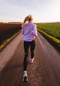 Athlete in a light purple long-sleeve top and black leggings with cut-outs, running on a dirt path beside fields at sunset.