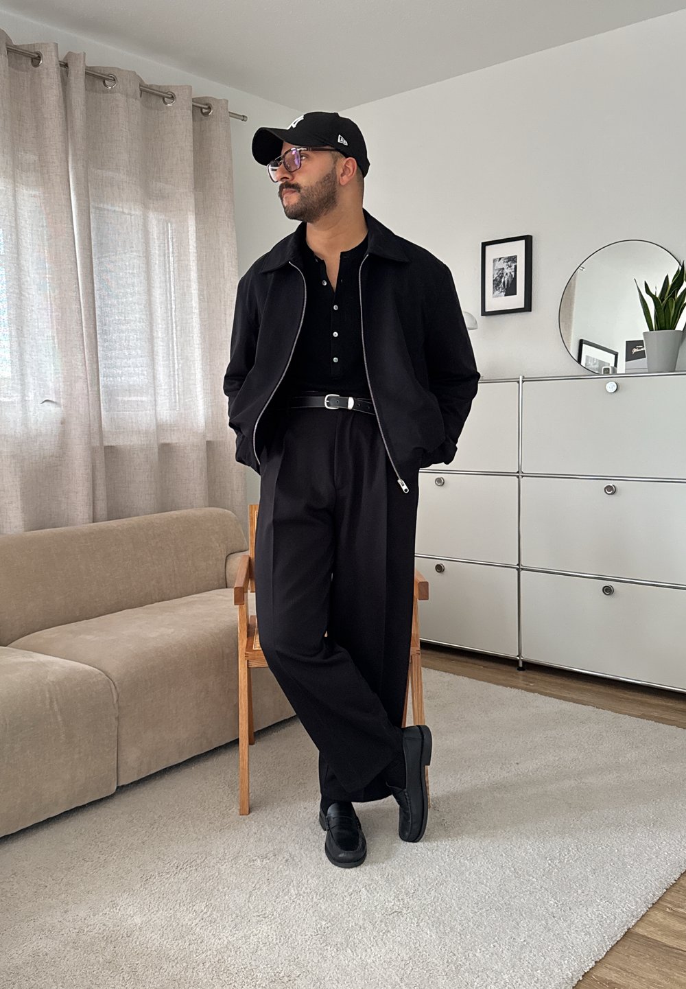 Svart jakke over en svart skjorte med krage, i kombinasjon med svarte bukser og sko. Inkluderer briller og en svart caps. Nøytral, moderne interiør.