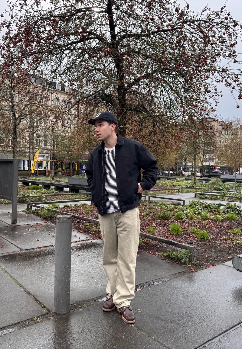 Homme portant une veste noire, une chemise rayée, un pantalon beige et une casquette, debout sur un trottoir mouillé près d'un arbre portant des baies rouges dans un parc urbain.