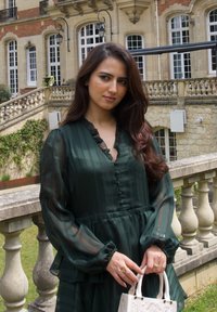 Vestido largo de color verde oscuro, de manga larga, con escote con volantes y tela texturizada. Presenta un bolso blanco y anillos. Edificio histórico al fondo.