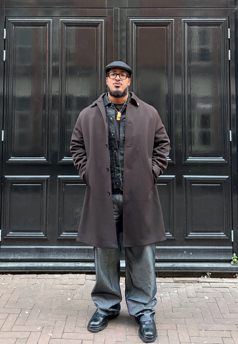 Homme avec lunettes et casquette noire portant un long manteau sombre, un pantalon gris et des chaussures noires, debout devant une grande porte noire à panneaux.