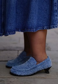 Άτομο που φορεί μπλε παπούτσια με χαμηλό τακούνι, με γούνα, και μπλε τζιν φούστα, στέκεται σε πέτρινο πλακόστρωτο.