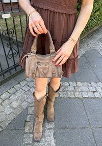 Asmuo, apsirengęs rudos spalvos suknele ir batais, laiko taupe spalvos zomšos rankinę su užtrauktuku, stovėdamas ant akmenų ir šaligatvio takelio.