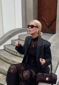 Crna kožna oversize blazerica preko crne prozirne čipkaste suknje, u kombinaciji s crnim topom, oversized sunčanim naočalama, zlatnim okruglim naušnicama i bordo torbom.